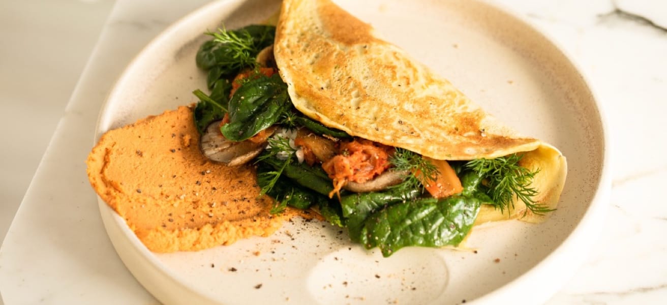 A bright photograph of a golden yellow omelette. It is served folded over a mix of vegetables on a stone coloured plate placed on a counter top