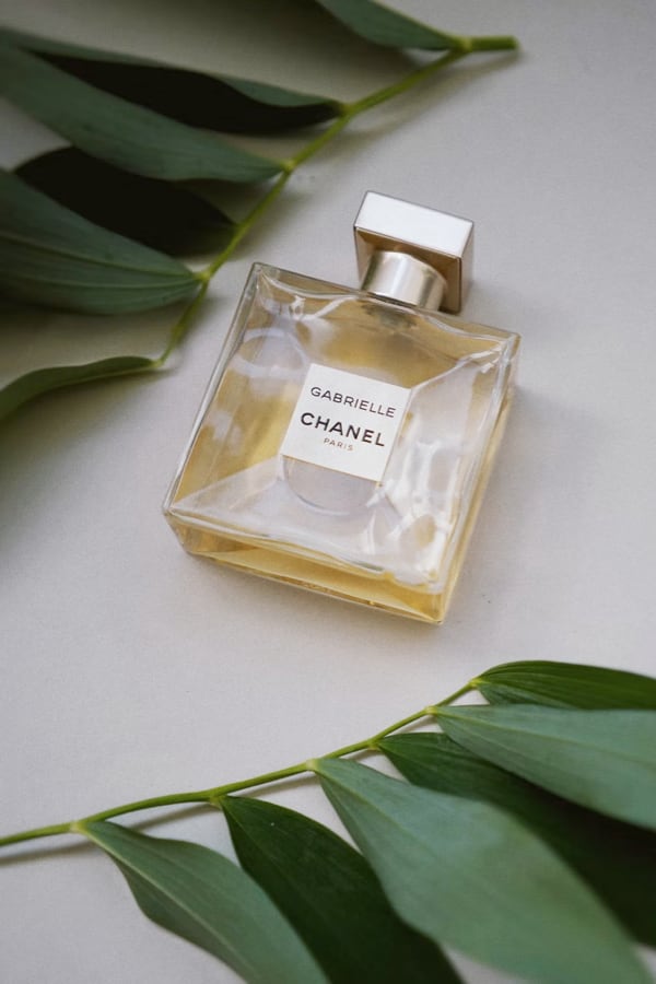 A wide photograph of a bottle of golden coloured perfume labelled Gabrielle Chanel Paris sitting on a table surrounded by green leafs